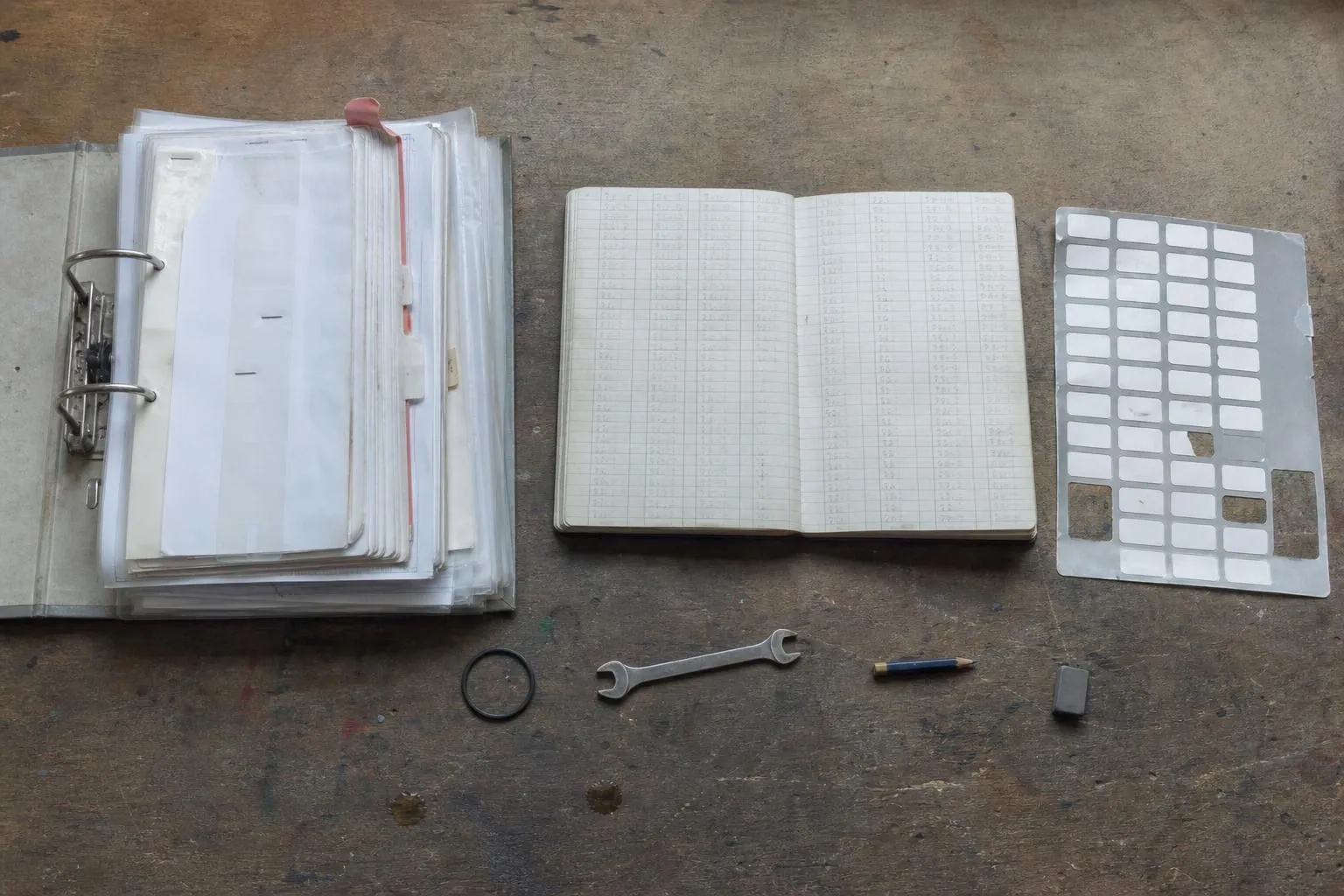 Vue de dessus d’un établi avec un classeur à anneaux ouvert et gondolé par l’humidité, un carnet à réglure et une planche d’étiquettes numérotées.
